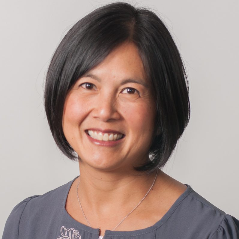 Leilani Yau smiling at the camera, wearing a gray top and a necklace, with a light gray background.